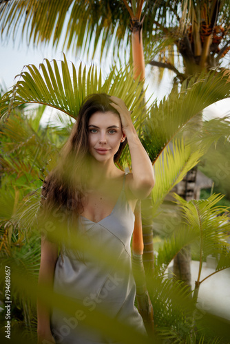 Fashion shot of young gorgeous female. stand near tropival palm tree outdoor, touch her hair and look at the camera