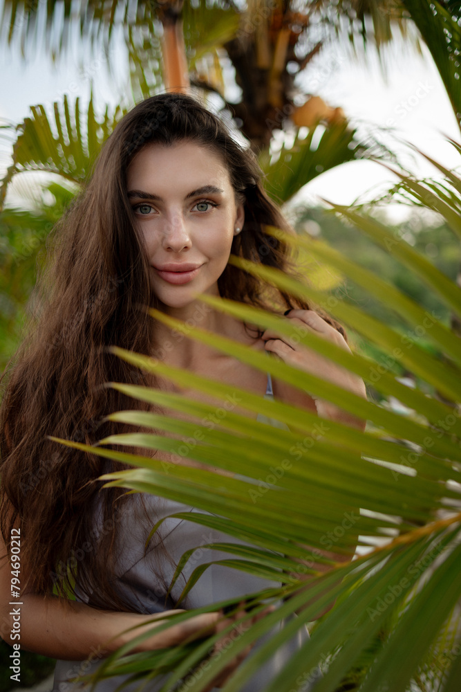 Close up face of beautiful young woman standing behind big green tropical palm leaves, looking at camera. Portrait of beauty woman with natural makeup Close up face of beautiful young woman standing behind big green tropical palm leaves, looking at camera. Portrait of beauty woman with natural makeup