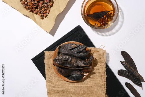 A dish of black locust fruits placed on sackcloth towel is spread on a rectangle black tray next to a glass bowl with brown liquid and some dried rose buds. Space for advertising or designing photo