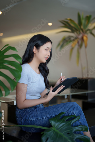 Asian teenage student woman watching video online lesson and writing notes on digital tablet while studying about education knowledge and doing assignment homework for preparing test exam in cafe