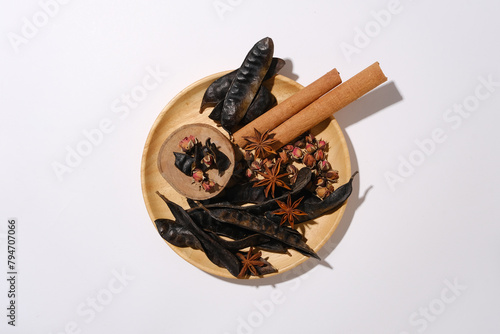 A round wooden dish placed in the center isolated on white table, contains some black locust pods, cinnamon, anise and dried rose buds. Layout suitable for cosmetic advertise  photo