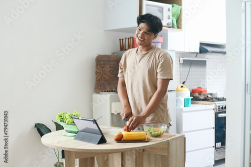 Young Man Making Salad Chopping Tomato while Looking at Online Tutorial on Device Tablet At The Kitchen 