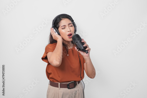 Overjoyed young woman wearing brown shirt and headphone to listen music and singing along isolated over white background. (ID: 794738810)