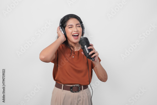 Overjoyed young woman wearing brown shirt and headphone to listen music and singing along isolated over white background. (ID: 794739022)