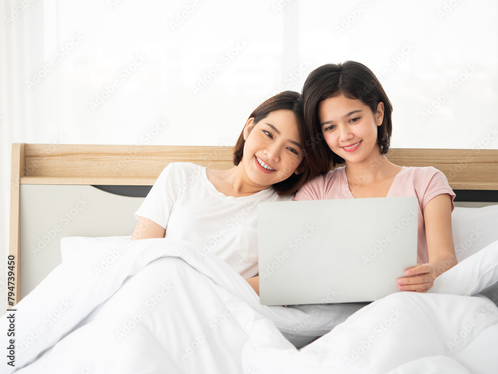 Asian two smiling women enjoying time together while looking at a laptop screen cozy and relaxed in a sunlit bedroom Asian two smiling women enjoying time together while looking at a laptop screen cozy and relaxed in a sunlit bedroom