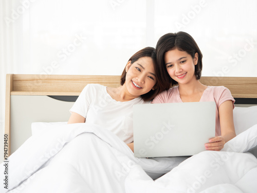 Asian two smiling women enjoying time together while looking at a laptop screen cozy and relaxed in a sunlit bedroom