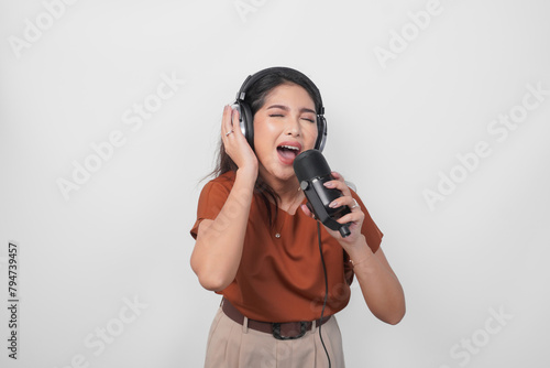 Overjoyed young woman wearing brown shirt and headphone to listen music and singing along isolated over white background. (ID: 794739457)