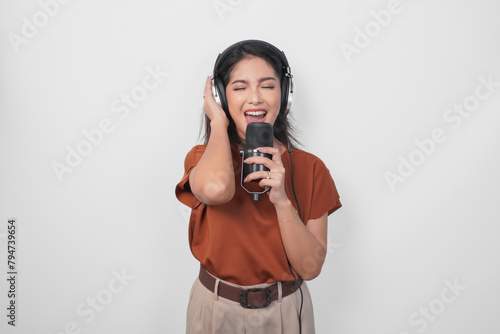 Excited young woman wearing brown shirt and headphone to listen music and singing along to the microphone isolated over white background. (ID: 794739654)