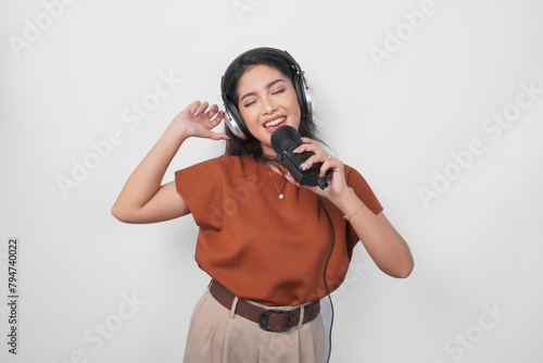 Excited young woman wearing brown shirt and headphone to listen music and singing along to the microphone isolated over white background. (ID: 794740022)