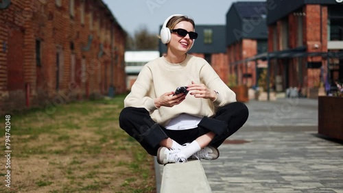 Woman in great mood having fun outdoors while listening to favorite songs and wearing headphones. Female holding smartphone and moving in rhythm of energetic song while sitting with crossed legs. photo