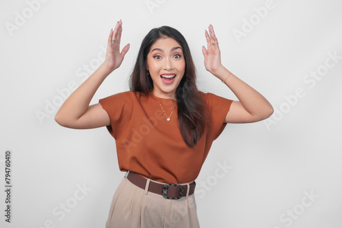 Excited and surprised young Asian woman wearing brown shirt standing isolated over white background. (ID: 794760410)
