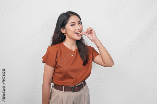 Beautiful young Asian woman in brown shirt holding and biting a medicine pill isolated by white background.