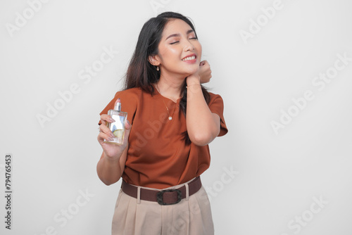 Beautiful young Asian woman in brown shirt holding bottle of perfume and spraying on her neck isolated by white background. (ID: 794765453)