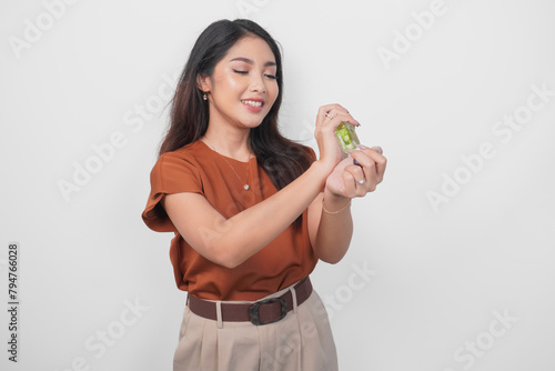 Smiling young Asian woman in brown shirt holding bottle of perfume and spraying on her wrist isolated by white background. (ID: 794766028)