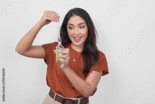 Happy young Asian woman in brown shirt holding bottle of serum to apply skin care isolated by white background. (ID: 794766244)