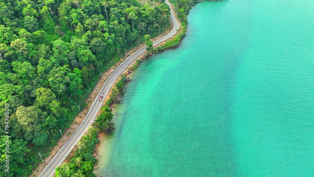 Aerial shot captures cars on a stunning tropical road, hugging the lush coastline, with vivid turquoise sea stretching endlessly. Travel and nature concept. Ko Chang, Thailand.

