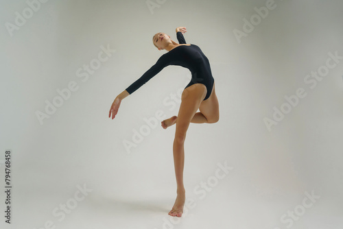 young ballerina in a black bodysuit shows ballet steps in motion standing on one leg and spreading her arms