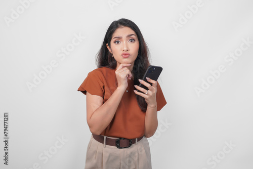 Thoughtful young woman wearing brown shirt looking aside while holding smartphone isolated over white background.