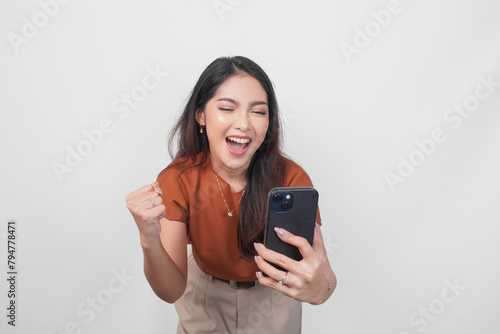 Joyful young Asian woman in brown shirt doing video call with happy successful expression and clenched fist gesture isolated over white background.