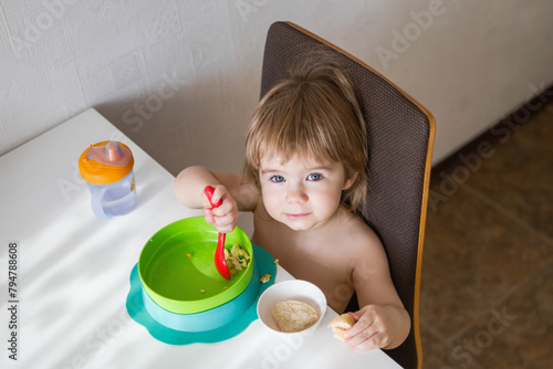 A hungry toddler sits in a high chair and eats breakfast. The child holds a spoon and eats from a green bowl. There is bread on the table. Isolated on a white background.