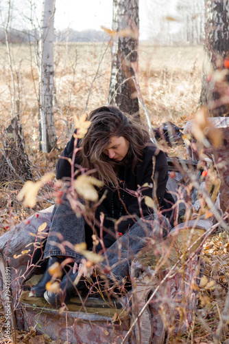 A young woman with long brown hair sits on the ground in a forest, eyes closed, smiling peacefully. She wears a black sweater and jeans. Tall grass and yellow leaves surround her.