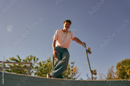 Low angle of happy male skateboarder holding skateboard standing at skate park in sunny day