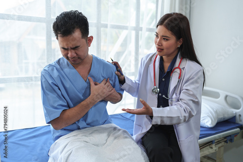 A professional female doctor in a white robe spoke. Has a heart rate stethoscope around his neck. Talk to a male patient about chest pain. Heart muscle to plan treatment at hospital bed.