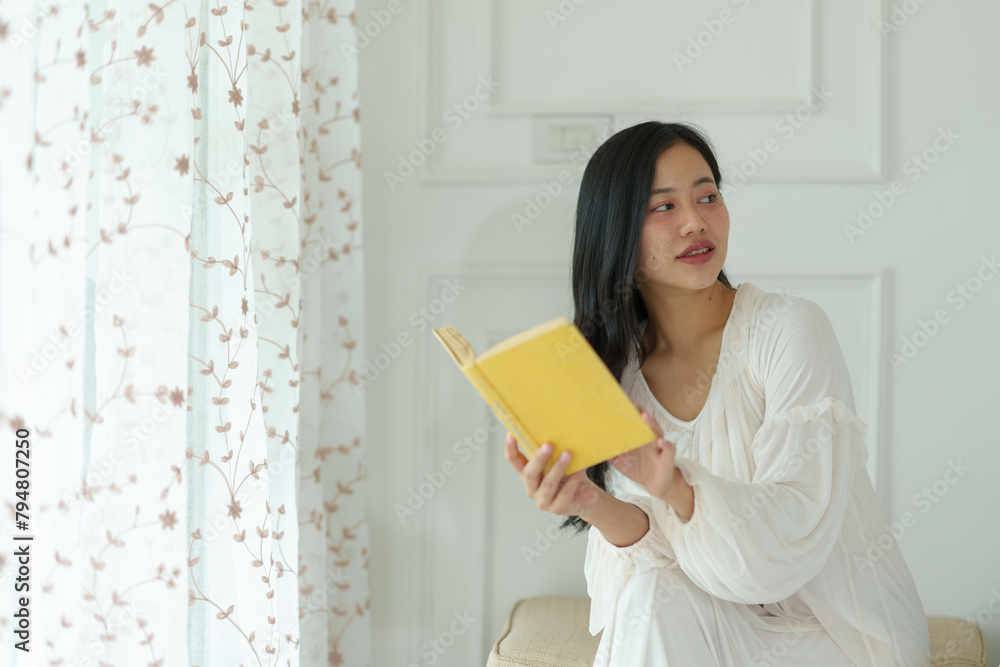 Asian woman in white pajamas sitting on the bed reading a book Ready to smile in her room in the morning, relaxing. Holiday concept, lifestyle. Asian woman in white pajamas sitting on the bed reading a book Ready to smile in her room in the morning, relaxing. Holiday concept, lifestyle.