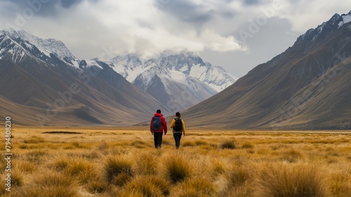 A backpacker couple pauses during their trek  admiring the panoramic view of a sprawling field against the backdrop of snow-dusted mountain ranges