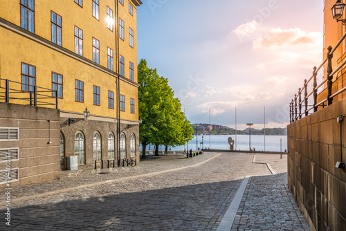 Warm sunlight bathes the historic buildings and cobblestone streets at Riddarholmen, overlooking the tranquil waterfront as day transitions to evening. Stockholm, Sweden