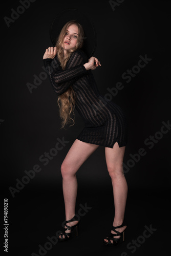 Breathtaking young woman striking confident pose with arms crossed at shoulder height on black background. Blonde with long hair and short knitted black dress, wide-brimmed felt hat, high heeled shoes