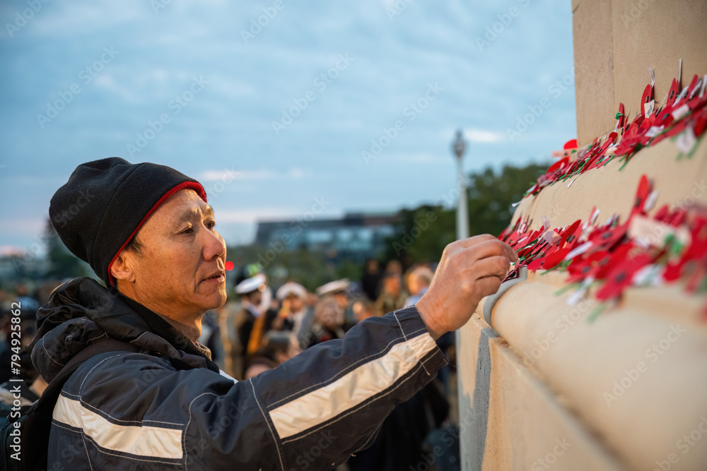 Man placing red poppies on the wall at the Court of Honour. Unrecognizable people in uniform in the background. Anzac Day commemoration. Auckland Domain.