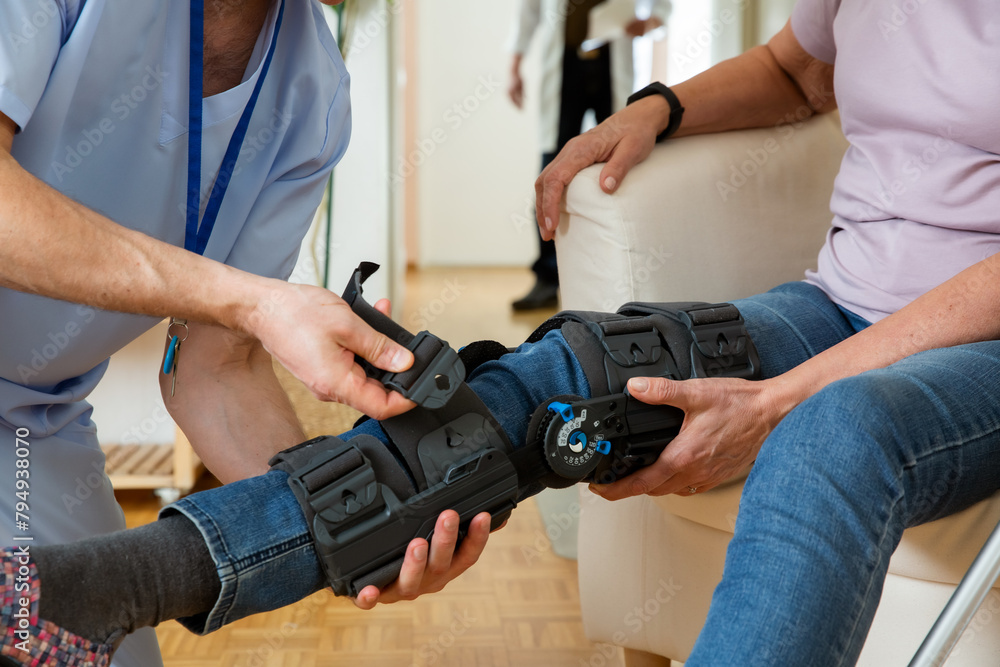 A male nurse helps to put a bondage, medical splint, knee brace on the leg of an elderly female patient who communicates with the attending physician at the clinic or hospital. A male nurse helps to put a bondage, medical splint, knee brace on the leg of an elderly female patient who communicates with the attending physician at the clinic or hospital.