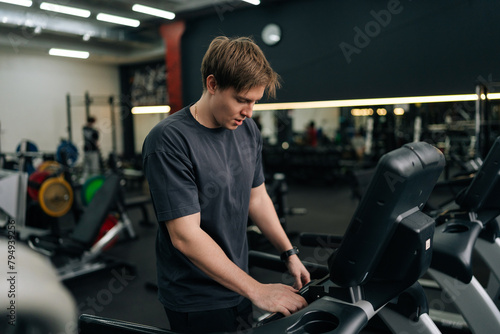 Portrait of athletic man in activewear adjusting settings on treadmill, preparing for cardio session in well-equipped gym. Sporty male walking on running track, working on health and fitness. © dikushin