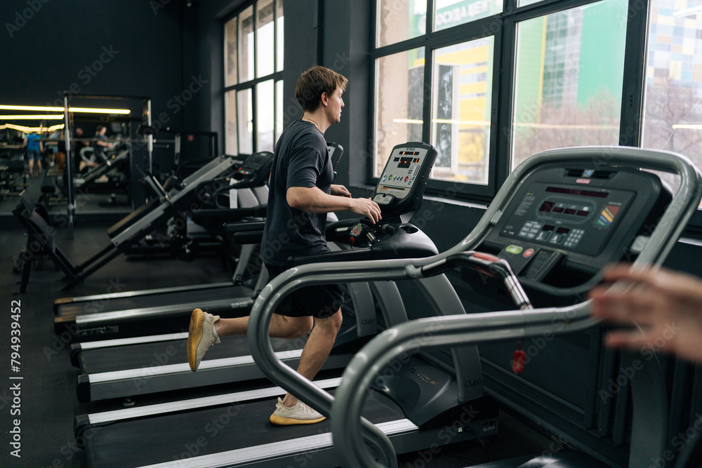 Rear remote view of athletic man jogging on gym machine, having cardio session in well-equipped fitness club. Side view of beginner sportsman having intensive cardio workout to burn calories in gym. Rear remote view of athletic man jogging on gym machine, having cardio session in well-equipped fitness club. Side view of beginner sportsman having intensive cardio workout to burn calories in gym.