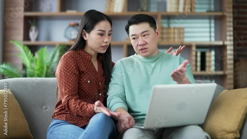 Asian family couple arguing while shopping online using laptop while sitting on sofa in living room at home. They are looking for a product in online store. A husband is unhappy with his wife's choice photo