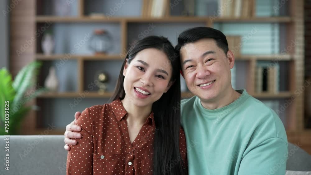 Close up portrait of happy asian married couple hugging while sitting on couch in living room looking at camera posing. Loving positive wife and husband hug and smile. Joyful moments of life together Close up portrait of happy asian married couple hugging while sitting on couch in living room looking at camera posing. Loving positive wife and husband hug and smile. Joyful moments of life together