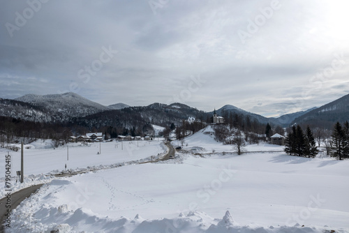 ampia visuale panoramica del piccolo paese di Lokve, in Slovenia, circondato da montagne, colline e vasti boschi completamente innevati, sotto un cielo coperto e grigio, in inverno photo