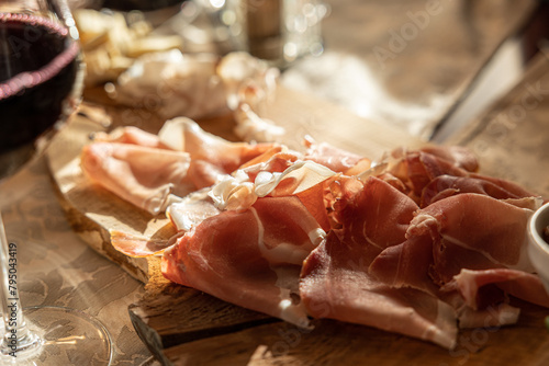 Obraz na płótnie primo piano su di un tagliere con dei salumi e affettati a base di carne, su un tavolo apparecchiato, vicino ad un calice di vino rosso, illuminati da dietro dalla luce del sole w salonie