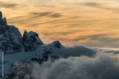 visuale dettagliata e da lontano di un'immensa quantità di nuvole basse che avanzano tra le valli in mezzo alle montagne innevate, in inverno, al tramonto, con un cielo colorato di arancione photo