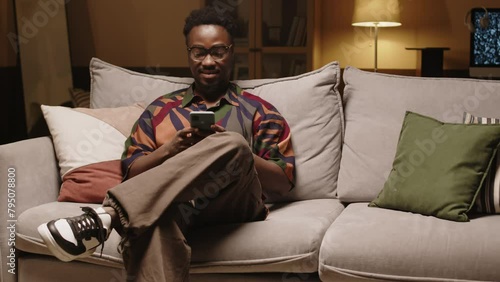 Medium shot of young Black hipster man in colorful printed shirt sitting on couch in modern living room and scrolling on smartphone in afternoon photo