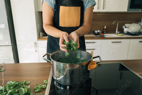 A woman is cooking greens in a pot on a stove, the pot is on the glass-ceramic hob