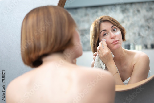 Attractive woman looking at mirror and applying moisturizing cream on cheeks in bathroom. Groomed young girl doing skincare morning routine after taking a shower