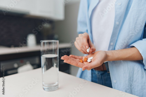 Woman holding pills and a glass of water on countertop in a modern kitchen interior with natural light