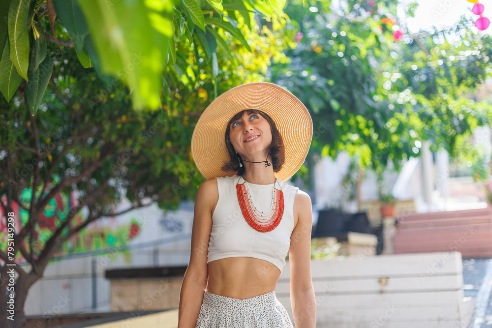 Fototapeta premium Happy beautiful fashionable woman with a smile wearing a fashionable hat. standing and having fun near a green tree on the street.