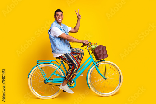 Full size photo of cheerful nice man dressed denim vest riding cycle to empty space show v-sign isolated on yellow color background
