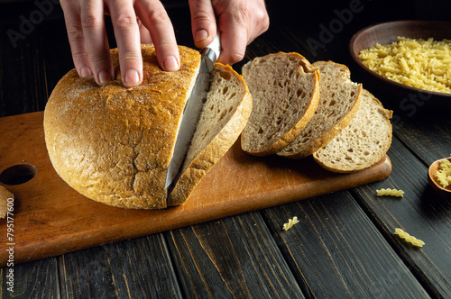 Chef hands cutting fresh bread with a knife on a kitchen board for lunch. Low key concept of healthy eating and traditional baked goods.