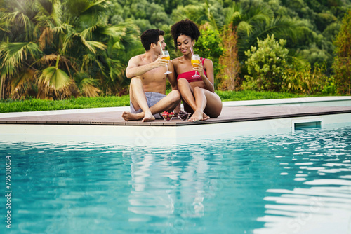 Multiethnic couple enjoys a romantic poolside date - sharing fruit juices cocktails and strawberries, surrounded by tropical greenery.