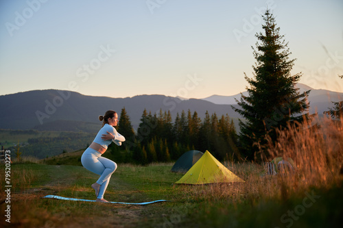 Woman practicing yoga outdoors in the mountains in a serene, natural setting near camping. Female performing yoga pose on mat, with backdrop of beautiful mountain landscape at sunrise or sunset.
