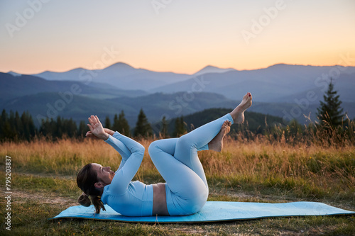 Woman practicing yoga outdoors in the mountains in a serene, natural setting. Female performing yoga pose on blue mat, with backdrop of beautiful mountain landscape at sunrise or sunset.